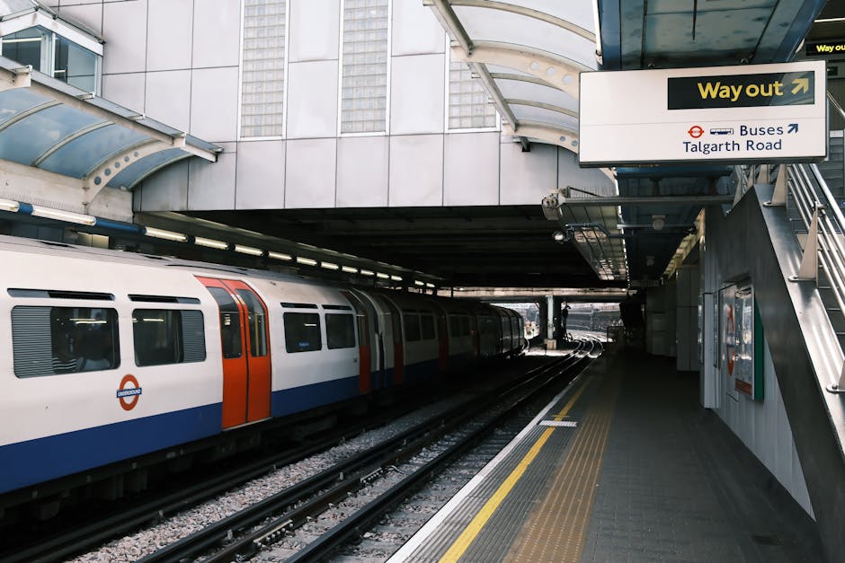 An underground train station platform at Hammersmith with a modern train parked on the tracks. The platform surface is clean and well-maintained, featuring textured yellow safety lines along the edge. Overhead, a curved metallic canopy provides shelter, supported by steel beams and illuminated by bright lighting. A prominent black and yellow 'Way out' sign with an arrow points to the station exit, with additional signage indicating directions to buses and Talgarth Road. The surrounding area includes glass and metal architectural elements, reflecting a sleek, urban design typical of city transit stations. Hammersmith Carpet Cleaning emphasizes surface cleaning and maintenance for public spaces in their deep cleaning and hygiene service solutions, ensuring cleanliness and safety for commuters and station staff.