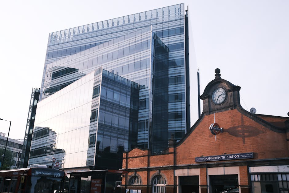 A modern glass office building with reflective windows towering over the historic brick Hammersmith station building, which features a clock on its facade. The station's sign indicating 'HAMMERSMITH STATION' is visible below the clock, with a nearby bus stop. The scene is captured during daylight with clear, bright weather, highlighting the contrast between the sleek, contemporary structure and the traditional brick architecture. Reflections of surrounding buildings are visible on the glass surfaces of the office tower, which is part of the urban landscape in Hammersmith. This image relates to surface cleaning and maintenance for commercial properties, emphasizing the importance of cleanliness in both modern and historic structures, and is representative of the services offered by Hammersmith Carpet Cleaning in addressing access problems and maintaining hygiene standards in such environments.