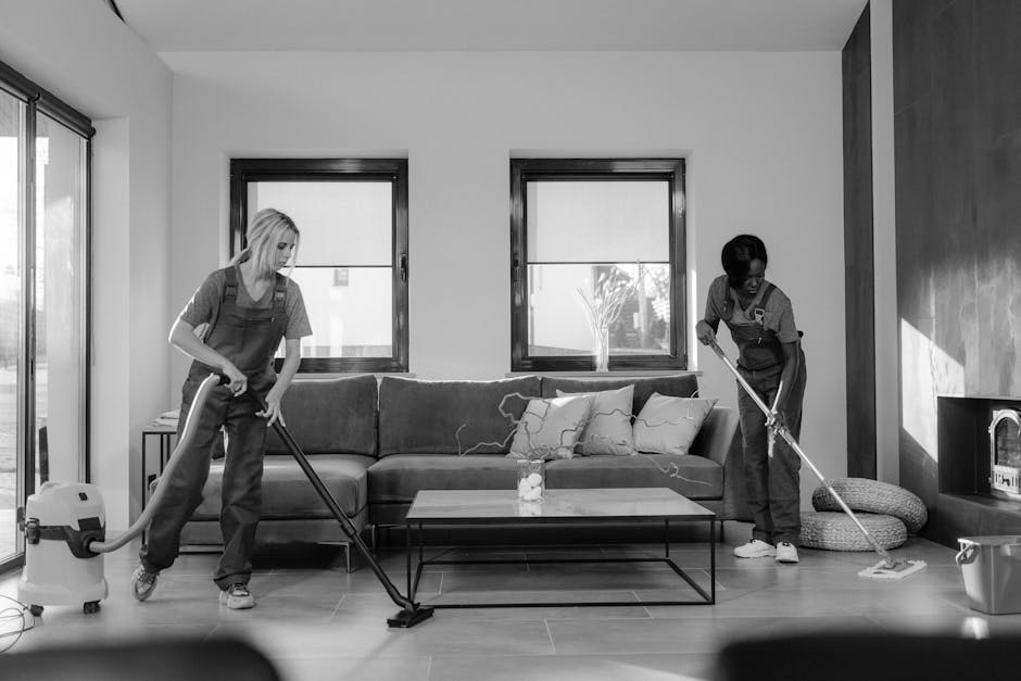 Two professional cleaners from Hammersmith Carpet Cleaning are performing deep surface cleaning in a modern living room, using vacuum cleaners and mops. The room features a light-colored wooden floor, a plush sofa with cushions, and large windows with black frames allowing natural light to illuminate the space. One cleaner is vacuuming the floor near the sofa, while the other is mopping the area closer to the TV stand, which holds a small decorative item. The room appears tidy, with a neutral color palette, and the cleaning tools are in use to ensure thorough sanitation of the floors and surfaces as part of a comprehensive domestic cleaning service. Bright natural light emphasizes the cleanliness and order of the room, supporting high standards of hygiene typical for end of tenancy cleaning preparations.