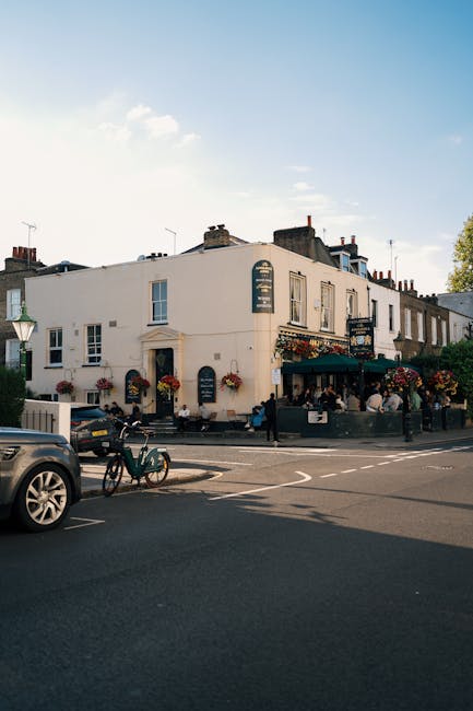 A street scene featuring a white, two-story building with multiple windows, outdoor seating areas, and hanging flower baskets. The building appears to be a café or pub, with patrons seated outside under green umbrellas. The pavement in front of the building is clean and well-maintained, with a bicycle parked near a lamppost and a car parked along the street. Bright daylight illuminates the scene, highlighting the building's exterior and the surrounding urban environment. Hammersmith Carpet Cleaning offers comprehensive surface cleaning and maintenance services, ensuring hygiene and cleanliness in both residential and commercial spaces. This image showcases a typical urban façade that may require regular cleaning to preserve its appearance and sanitation.