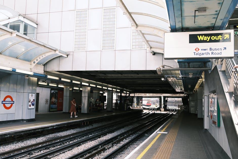 The image depicts a modern underground train station platform with concrete flooring and metal rail tracks extending into the distance. The platform features white tiled walls with signage, including a prominent roundel logo for Hammersmith, and directional signs indicating the way out to buses and Talgarth Road. A person dressed in casual clothing is walking along the platform, and there are waiting areas with benches and posters visible on the walls. Overhead, a large curved glass and metal canopy provides shelter, with bright lighting illuminating the area. The cleanliness of the station appears well-maintained, with no visible dirt or litter, and the surfaces of the floor and tracks are free of debris, reflecting effective cleaning by professionals such as Hammersmith Carpet Cleaning for maintaining hygiene standards in public transportation environments.