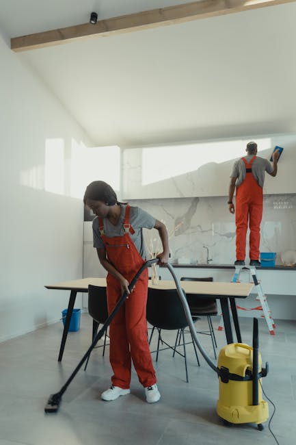 Two professional cleaners wearing red overalls and grey t-shirts are performing surface cleaning inside a modern kitchen. The cleaner in the foreground is vacuuming the gray tiled floor with a yellow portable vacuum cleaner, focusing on a wooden dining table with black chairs. The second cleaner, standing on a small step ladder, is wiping or polishing the glass backsplash behind the sink area. The kitchen features a sleek, white wall with a marble-patterned countertop and a well-lit, minimalist design. Natural light streams through large windows, creating a bright and airy environment. The scene highlights thorough cleaning and sanitisation processes typical of domestic cleaning services, and reflects the professional standards maintained by Hammersmith Carpet Cleaning.