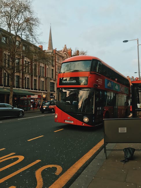 A double-decker bus with a red exterior and black accents, displaying the destination 'Hammersmith,' is driving along a street in Hammersmith W6. The street features paved sidewalks with a black bench and a small bag placed on it. Modern multi-story buildings with large windows and a mix of brick and stone facades line the street, while a church with a tall spire is visible in the background under an overcast sky. Road markings, including a yellow bus stop line, are clearly visible on the asphalt road, which is occupied by a few cars and pedestrians. The scene captures a typical urban street view in Hammersmith, emphasizing the area's transportation and architectural features. This image is used to illustrate the area surrounding the services offered by Hammersmith Carpet Cleaning as part of their cleaning and maintenance content.