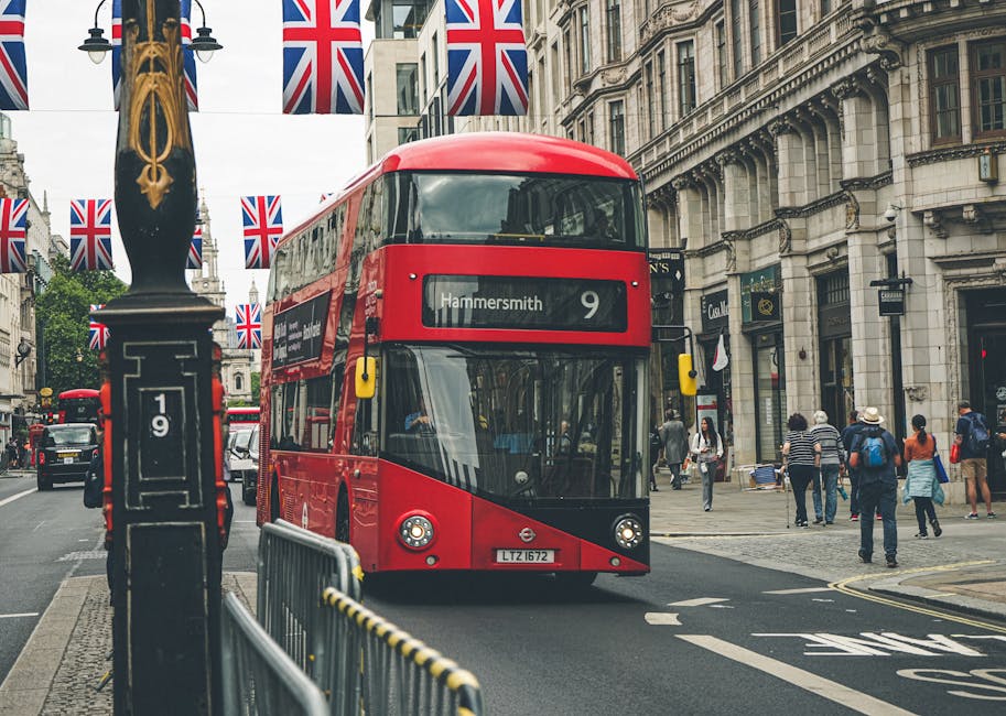 A red double-decker bus displaying 'Hammersmith 9' traverses a city street in Hammersmith, W6, with pedestrians walking on the sidewalk. The street is lined with historic stone buildings featuring ornate architecture and large windows. British Union Jack flags hang from lamp posts along the street, which is partly blocked by a metal barrier and a black lamppost with gold decorative elements. The scene is well-lit by natural daylight, with no visible dirt or clutter, showcasing a clean and orderly urban environment. This setting reflects an active, bustling commercial area as seen in the King Street shop cleaning Hammersmith W6 area, maintained by Hammersmith Carpet Cleaning.