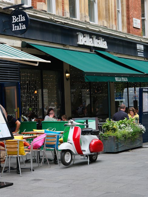 A double-decker bus with a red exterior and black accents, displaying the destination 'Hammersmith,' is driving along a street in Hammersmith W6. The street features paved sidewalks with a black bench and a small bag placed on it. Modern multi-story buildings with large windows and a mix of brick and stone facades line the street, while a church with a tall spire is visible in the background under an overcast sky. Road markings, including a yellow bus stop line, are clearly visible on the asphalt road, which is occupied by a few cars and pedestrians. The scene captures a typical urban street view in Hammersmith, emphasizing the area's transportation and architectural features. This image is used to illustrate the area surrounding the services offered by Hammersmith Carpet Cleaning as part of their cleaning and maintenance content.