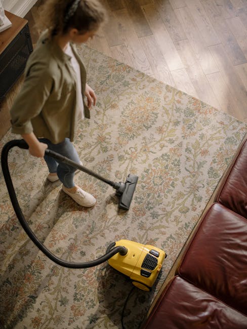 A woman engaged in surface cleaning using a yellow vacuum cleaner with a black hose, vacuuming a large, intricate-patterned rug with floral motifs in shades of beige, green, and orange. She is standing on a wooden floor with a natural finish, next to a red leather sofa visible at the edge of the frame. The room is well-lit, and the rug appears clean and dust-free, indicating a thorough deep cleaning process carried out by Hammersmith Carpet Cleaning as part of their domestic cleaning services.