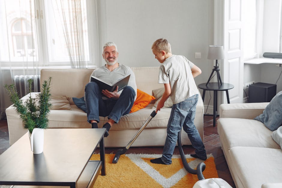 A young boy is vacuuming the carpet in a bright, modern living room while a smiling older man, possibly a grandfather, is sitting on a beige sofa reading a book. The room has large windows letting in natural light, white walls, and minimal decor including a small potted plant on a white side table. The carpet features a colorful geometric pattern in yellow, white, and orange tones. A black side table with a lamp and some cushions are visible in the background. The scene depicts routine domestic cleaning with a focus on surface cleaning and maintenance, with the Hammersmith Carpet Cleaning company image emphasizing hygienic practices for deep cleaning and sanitisation.